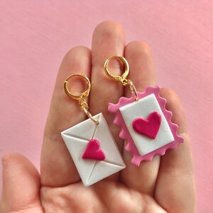 May include: A pair of gold hoop earrings with pink and white clay charms shaped like envelopes with pink hearts.