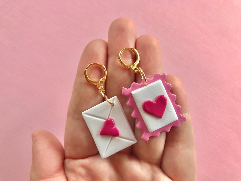 May include: A pair of gold hoop earrings with pink and white clay charms shaped like envelopes with pink hearts.