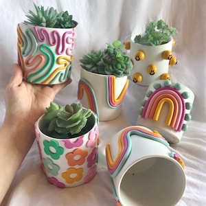 May include: Five white ceramic plant pots with different colourful clay designs. The pots are decorated with swirls, rainbows, flowers, and smiley faces. Each pot has a small succulent plant inside.