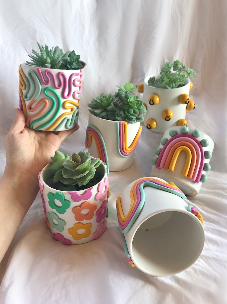 May include: Five white ceramic plant pots with different colourful clay designs. The pots are decorated with swirls, rainbows, flowers, and smiley faces. Each pot has a small succulent plant inside.