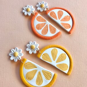 Puede incluir: Dos pares de pendientes, uno naranja y otro amarillo, con forma de rodajas de c&iacute;tricos. Cada pendiente presenta un centro blanco con segmentos naranjas o amarillos, una c&aacute;scara coloreada y un perno de margarita. El fondo es rosa claro.