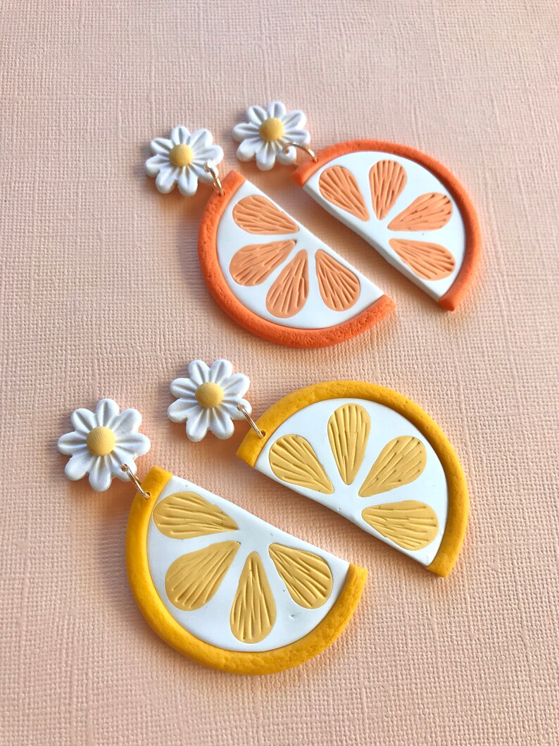 Puede incluir: Dos pares de pendientes, uno naranja y otro amarillo, con forma de rodajas de c&iacute;tricos. Cada pendiente presenta un centro blanco con segmentos naranjas o amarillos, una c&aacute;scara coloreada y un perno de margarita. El fondo es rosa claro.