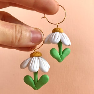 May include: A pair of gold hoop earrings with white and green polymer clay daisy flower charms. The flowers have yellow centers and green leaves.