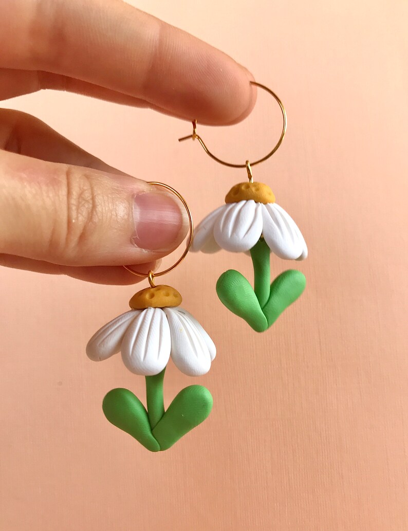 May include: A pair of gold hoop earrings with white and green polymer clay daisy flower charms. The flowers have yellow centers and green leaves.