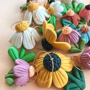 May include: A set of handmade polymer clay earrings featuring various flower designs. The earrings are made with white, yellow, green, purple, and brown clay. Each earring has a gold hoop.