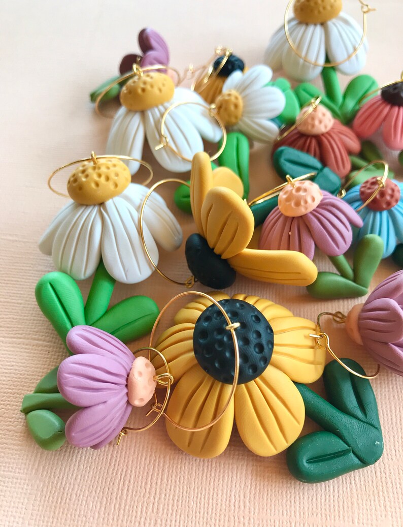 May include: A set of handmade polymer clay earrings featuring various flower designs. The earrings are made with white, yellow, green, purple, and brown clay. Each earring has a gold hoop.