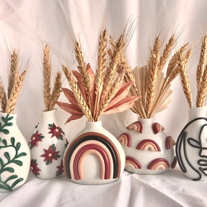 Pu&ograve; includere: Cinque vasi in ceramica bianca con diversi design, ciascuna con un mazzo di steli di grano secchi. I vasi presentano una vite verde, fiori rossi, un arcobaleno, forme di arcobaleno astratte e un disegno a linea nera di un viso.