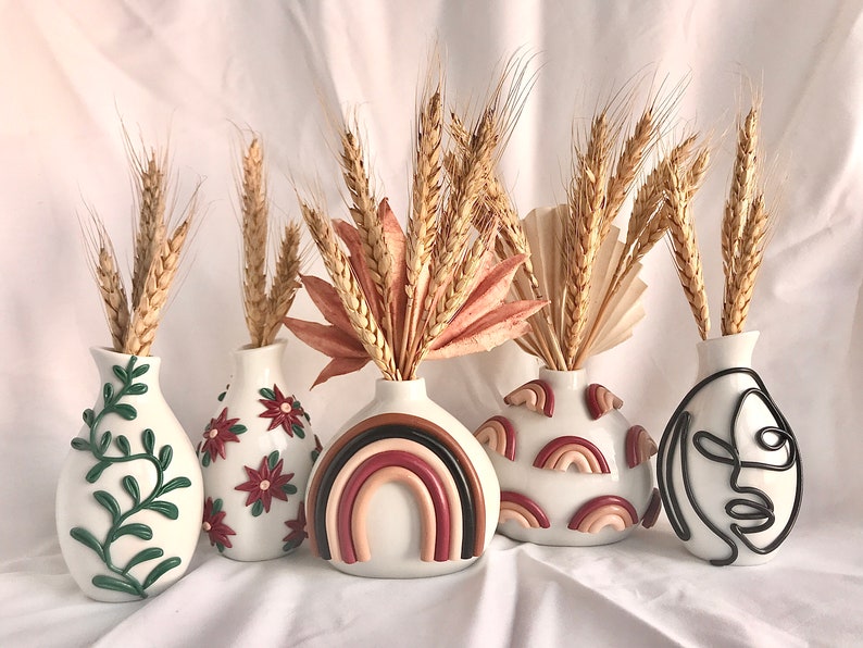 Pu&ograve; includere: Cinque vasi in ceramica bianca con diversi design, ciascuna con un mazzo di steli di grano secchi. I vasi presentano una vite verde, fiori rossi, un arcobaleno, forme di arcobaleno astratte e un disegno a linea nera di un viso.