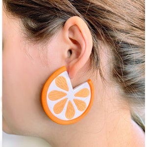 May include: Orange slice shaped earrings with a white center and orange edges. The earrings are made of a lightweight material and are perfect for adding a touch of whimsy to any outfit.