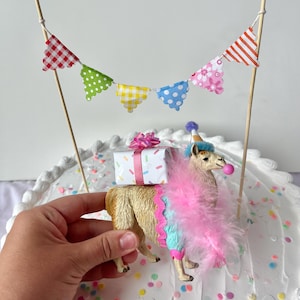 K&ouml;nnte beinhalten: Ein Lama-Kuchenaufsatz mit einer rosa Federboa, einer Partyh&uuml;tchen und einer Geschenkbox. Das Lama ist mit einer rosa und blauen Bord&uuml;re verziert. Ein farbenfrohes Banner ist &uuml;ber dem Lama aufgeh&auml;ngt. Der Kuchen ist mit Streuseln verziert.