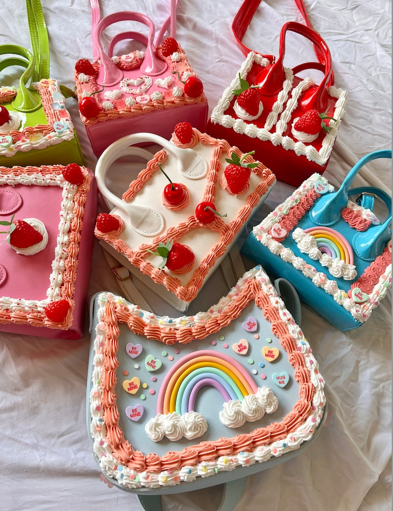 May include: A collection of colorful cake-themed handbags with various designs, including strawberries, cherries, rainbows, and frosting. The bags are made of a glossy material and have a variety of colors, including pink, red, white, blue, and green.