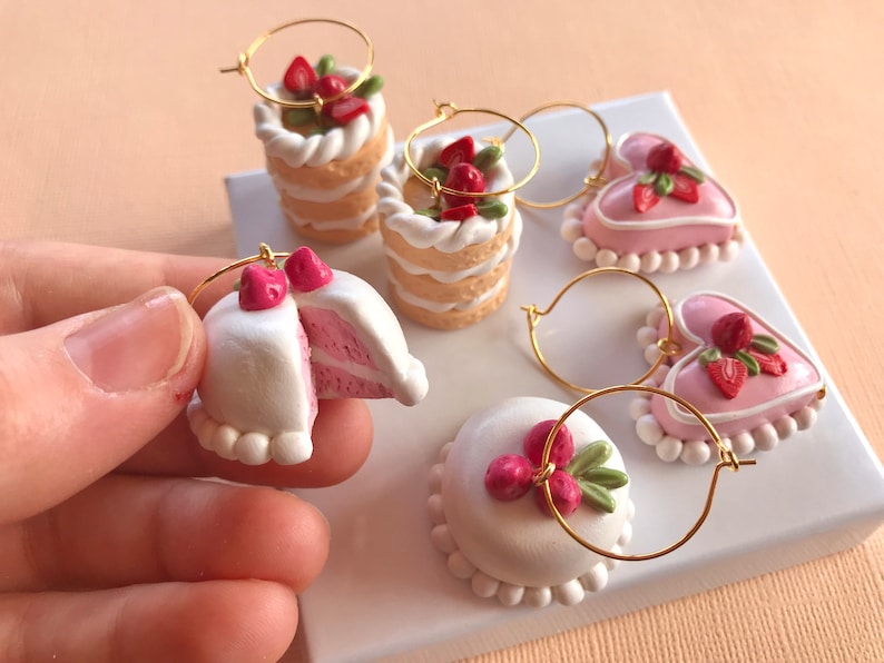 May include: A set of six handmade polymer clay earrings featuring miniature cakes and hearts decorated with pink frosting and red strawberries. The earrings are attached to gold hoops.