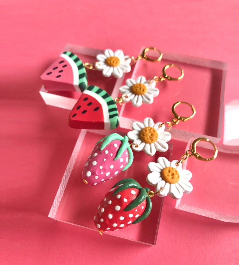 May include: A pair of earrings featuring a watermelon and a strawberry charm, each with a white daisy charm. The charms are attached to gold hoop earrings.