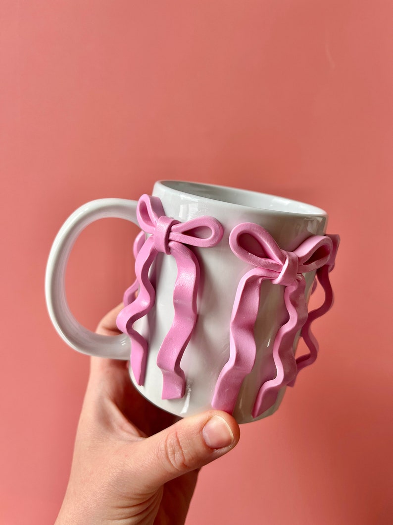 Pu&ograve; includere: Tazza in ceramica bianca con dettagli a nastro rosa. Il nastro &egrave; scolpito e ha una texture ondulata.