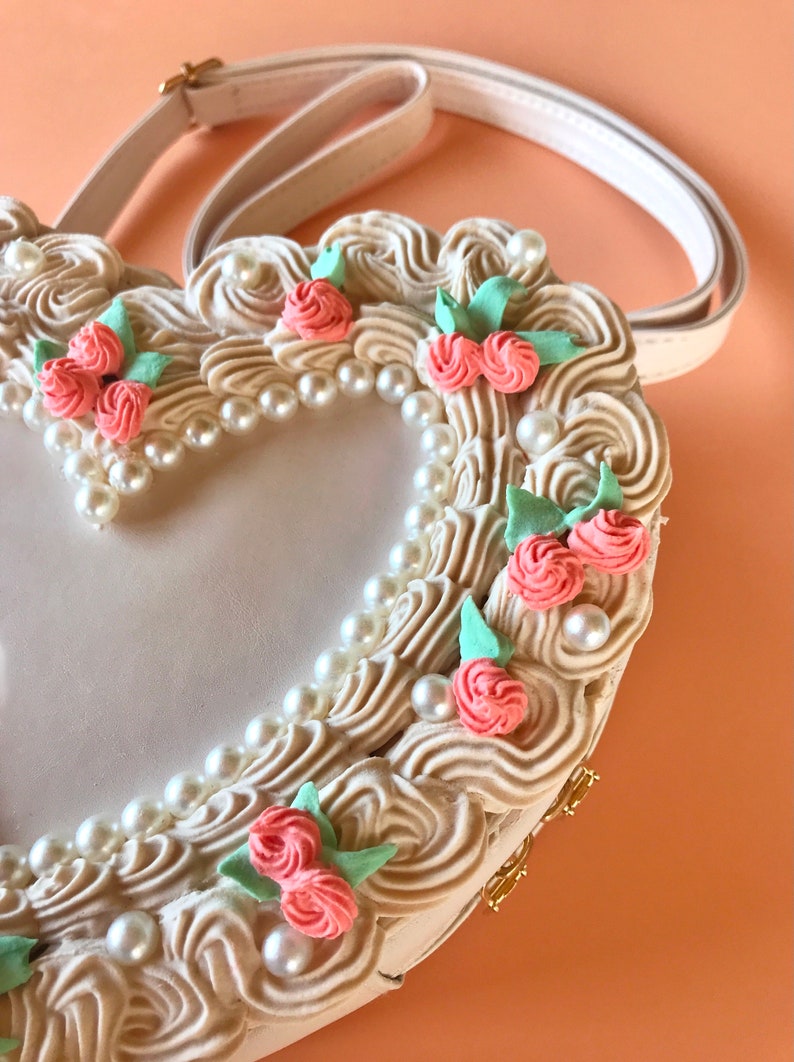 May include: A white heart-shaped handbag with a cream-colored, swirl-like design. The bag is decorated with faux pearls and pink flowers.