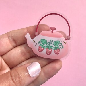 May include: A clear sticker of a pink teapot with a red handle and lid. The teapot is decorated with green leaves and three red strawberries. There are two small yellow bees flying around the teapot.