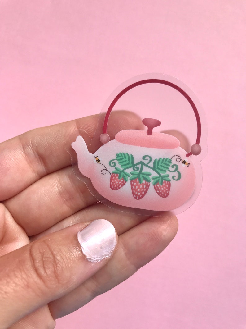 May include: A clear sticker of a pink teapot with a red handle and lid. The teapot is decorated with green leaves and three red strawberries. There are two small yellow bees flying around the teapot.