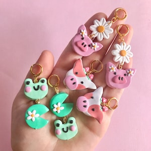 Pu&ograve; includere: Un set di orecchini fatti a mano in pasta polimerica con vari disegni di animali e fiori. Gli orecchini sono rosa, verdi e bianchi con ganci dorati. I disegni includono una rana, una margherita, un maiale e una mucca.