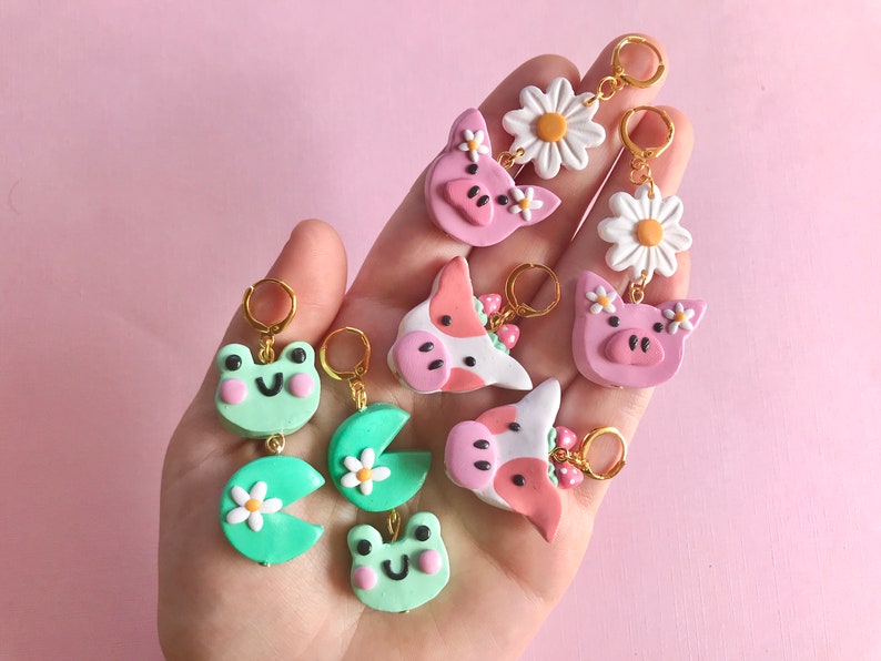 Pu&ograve; includere: Un set di orecchini fatti a mano in pasta polimerica con vari disegni di animali e fiori. Gli orecchini sono rosa, verdi e bianchi con ganci dorati. I disegni includono una rana, una margherita, un maiale e una mucca.