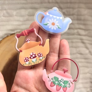 May include: Three clear vinyl stickers featuring teapots with different designs. The first teapot is blue with a white daisy, the second is yellow with red mushrooms, and the third is pink with red strawberries.