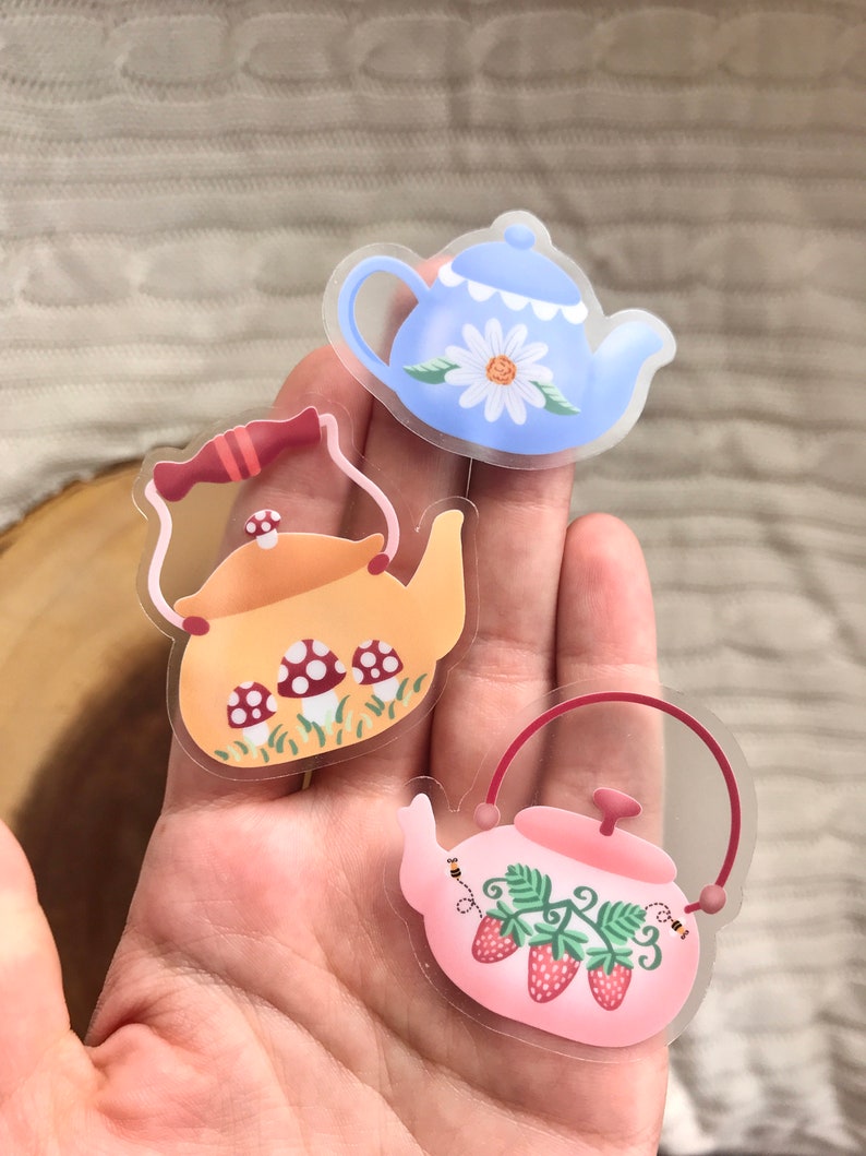 May include: Three clear vinyl stickers featuring teapots with different designs. The first teapot is blue with a white daisy, the second is yellow with red mushrooms, and the third is pink with red strawberries.