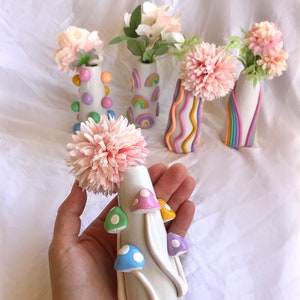 May include: Five ceramic vases with different designs. The vases are white with colorful details. The designs include polka dots, rainbows, wavy lines, and mushrooms. The vases are filled with pink flowers.