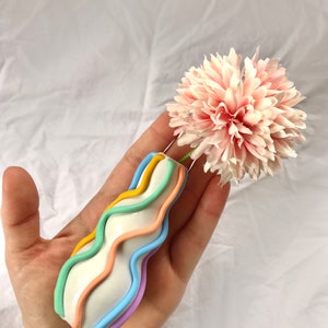 May include: A white ceramic vase with a wavy design and pastel colored stripes. The vase is holding a single pink flower.