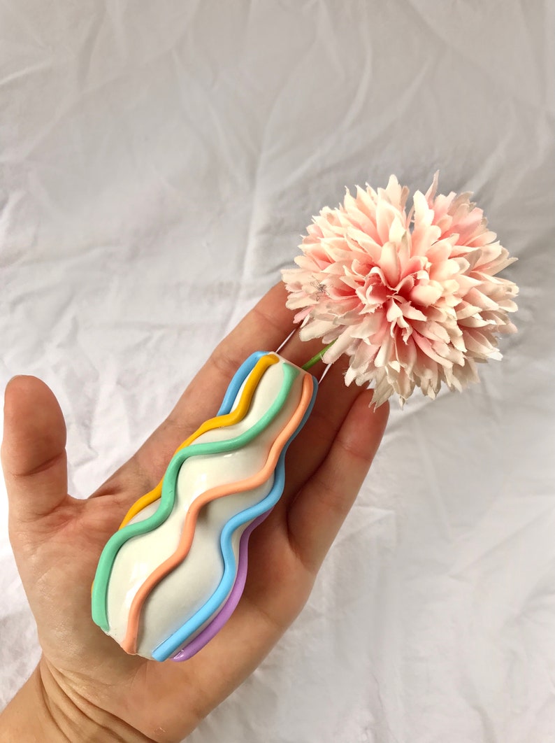 May include: A white ceramic vase with a wavy design and pastel colored stripes. The vase is holding a single pink flower.
