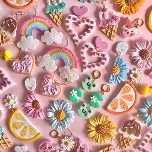 May include: A collection of colorful polymer clay earrings featuring various themes, including rainbows, hearts, ice cream cones, sunflowers, clouds, lemons, and frogs. The earrings are displayed on a pink background.