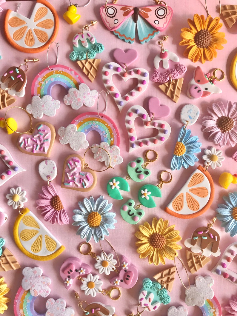 May include: A collection of colorful polymer clay earrings featuring various themes, including rainbows, hearts, ice cream cones, sunflowers, clouds, lemons, and frogs. The earrings are displayed on a pink background.