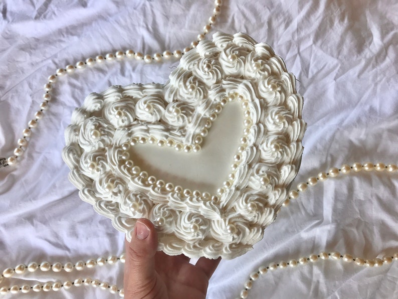 May include: A white heart-shaped box with a raised design of swirls and pearls around the edges. The box has a smaller heart-shaped opening in the center.