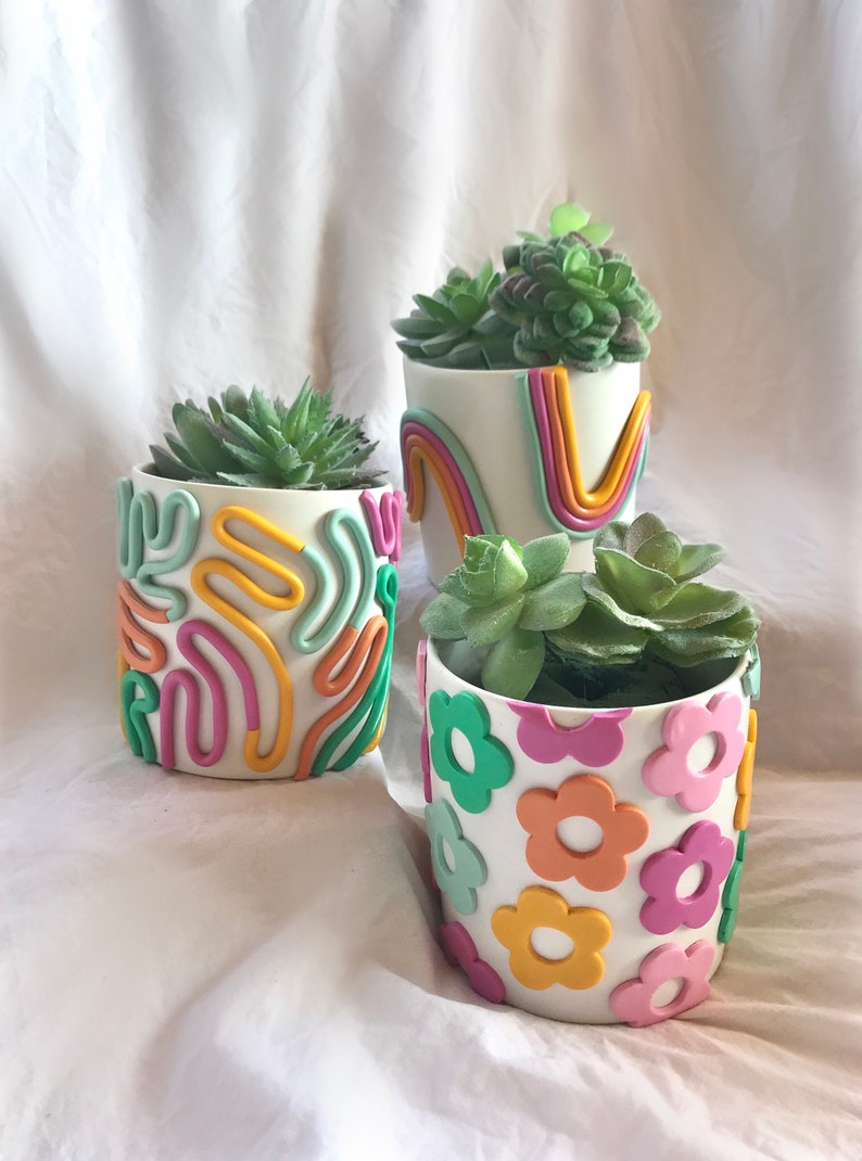 May include: Three white ceramic succulent planters with different colourful designs. The planter on the left has a pink, yellow, and green abstract design. The planter in the middle has a rainbow design. The planter on the right has a flower design with pink, yellow, and green flowers.