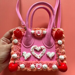 May include: Pink faux leather handbag shaped like a cake with white frosting, red cherries, and pink candy hearts that say "Be Mine".