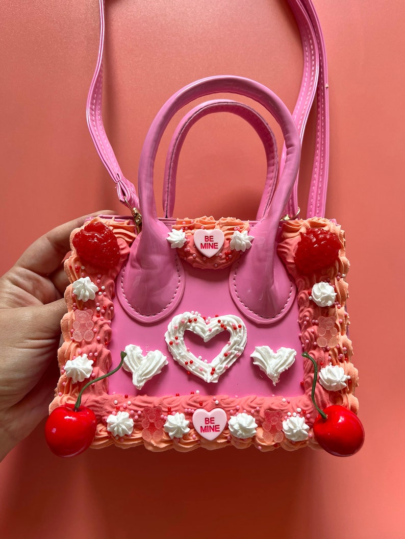 May include: Pink faux leather handbag shaped like a cake with white frosting, red cherries, and pink candy hearts that say "Be Mine".