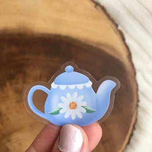 May include: A clear sticker with a blue teapot with a white daisy design. The teapot has a white scalloped rim and green leaves around the daisy.