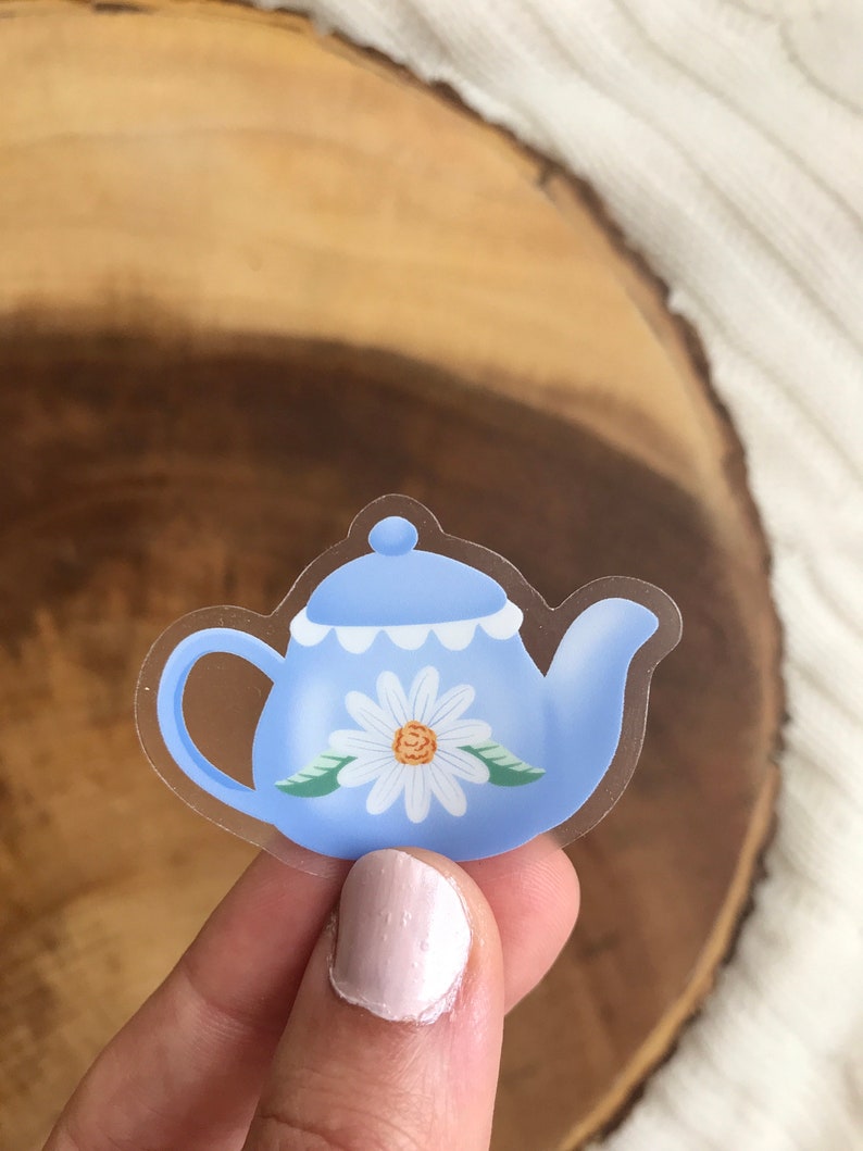 May include: A clear sticker with a blue teapot with a white daisy design. The teapot has a white scalloped rim and green leaves around the daisy.