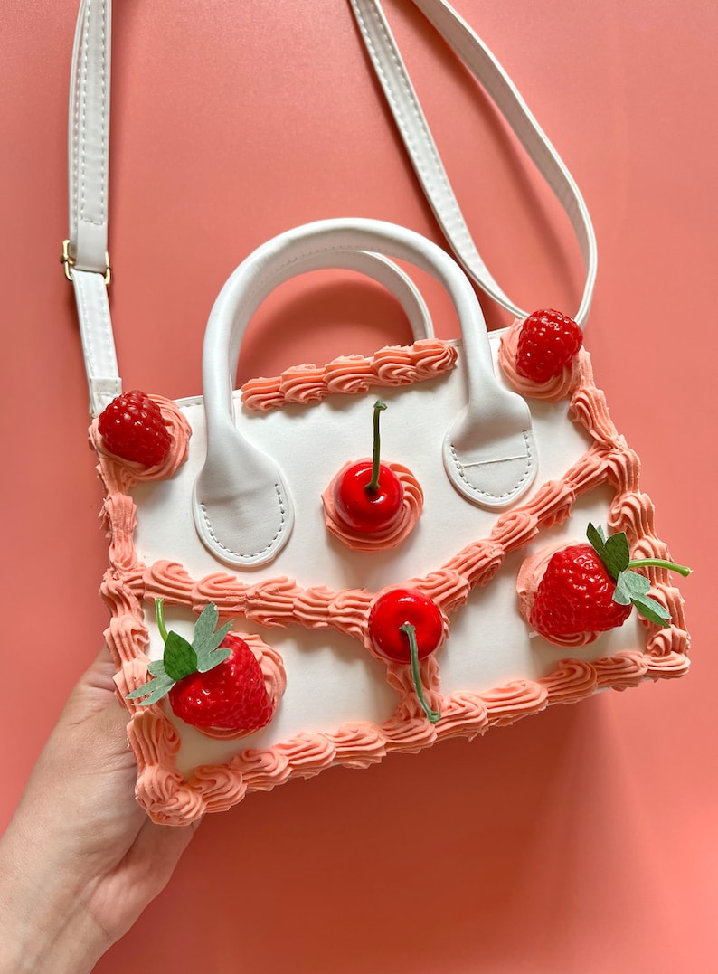 May include: A white handbag shaped like a cake with pink frosting and red cherries and strawberries. The bag has a white strap and a white handle.