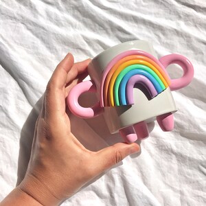 May include: A white ceramic mug with a rainbow design and pink handles. The mug has three pink feet.
