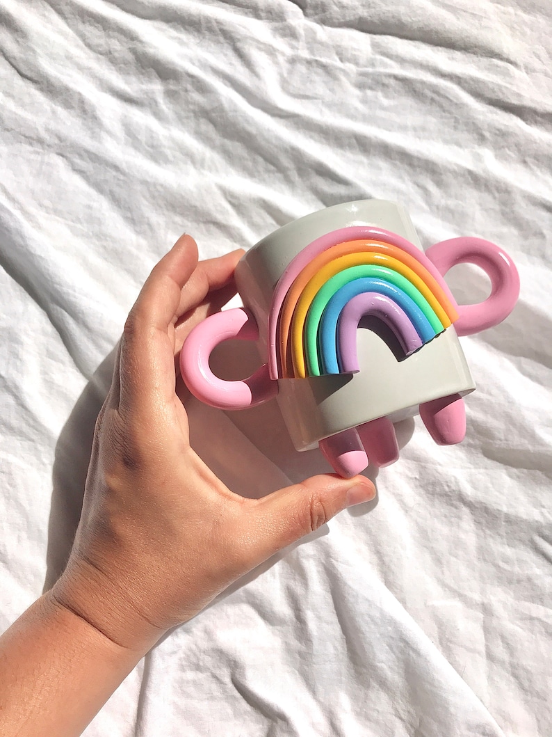 May include: A white ceramic mug with a rainbow design and pink handles. The mug has three pink feet.
