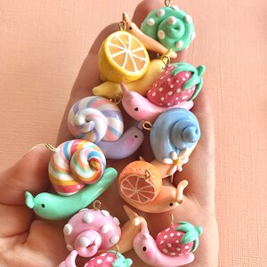 Pu&ograve; includere: Una collezione di ciondoli a forma di lumaca in pasta polimerica colorata con vari disegni, tra cui un limone, una fragola, un'arancia e una caramella a bastoncino. Le lumache sono tutte di colori diversi e hanno uno stile carino e fantasioso.
