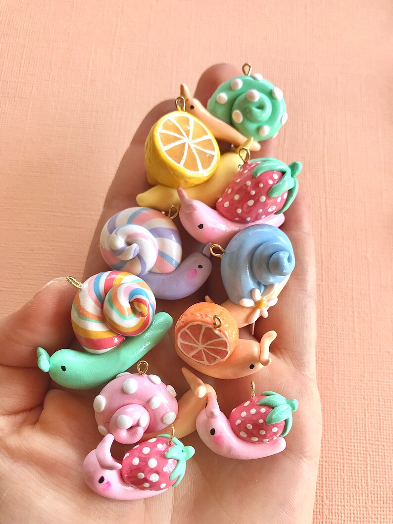 Pu&ograve; includere: Una collezione di ciondoli a forma di lumaca in pasta polimerica colorata con vari disegni, tra cui un limone, una fragola, un'arancia e una caramella a bastoncino. Le lumache sono tutte di colori diversi e hanno uno stile carino e fantasioso.