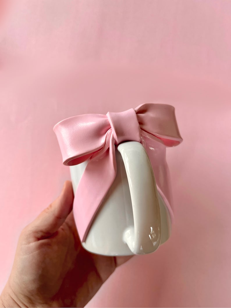 Pu&ograve; includere: Una tazza in ceramica bianca con un grande fiocco rosa legato attorno al manico. La tazza &egrave; tenuta in una mano.