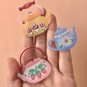 May include: Three clear teapots with different designs. The first teapot is yellow with red and white mushrooms. The second teapot is blue with a white daisy. The third teapot is pink with green leaves and red strawberries.
