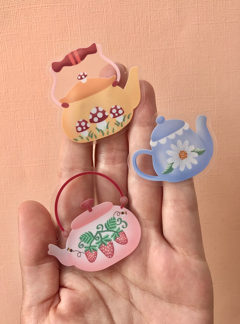 May include: Three clear teapots with different designs. The first teapot is yellow with red and white mushrooms. The second teapot is blue with a white daisy. The third teapot is pink with green leaves and red strawberries.