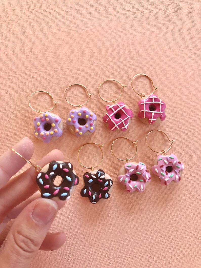 May include: Six pairs of donut earrings with gold hoops. The donuts are made of polymer clay and have different colored glazes and sprinkles. The donuts are pink, purple, and brown.