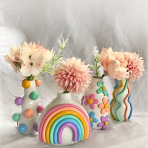 May include: Four colorful ceramic vases with pastel rainbow, flower, and polka dot designs. The vases are filled with pink and white flowers.