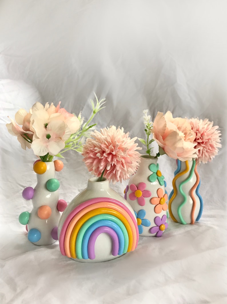 May include: Four colorful ceramic vases with pastel rainbow, flower, and polka dot designs. The vases are filled with pink and white flowers.