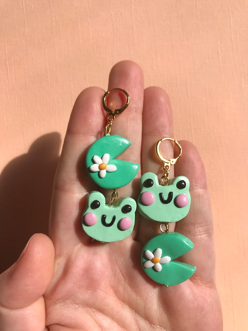 Pu&ograve; includere: Un paio di orecchini a forma di rana verde e Pac-Man verde con margherite bianche. Gli orecchini sono realizzati in pasta polimerica e hanno cerchi dorati.