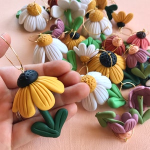 May include: A collection of handmade polymer clay flower earrings. The earrings feature a variety of flower designs, including daisies, sunflowers, and other wildflowers. The flowers are all hand-painted with vibrant colors and have a realistic appearance.