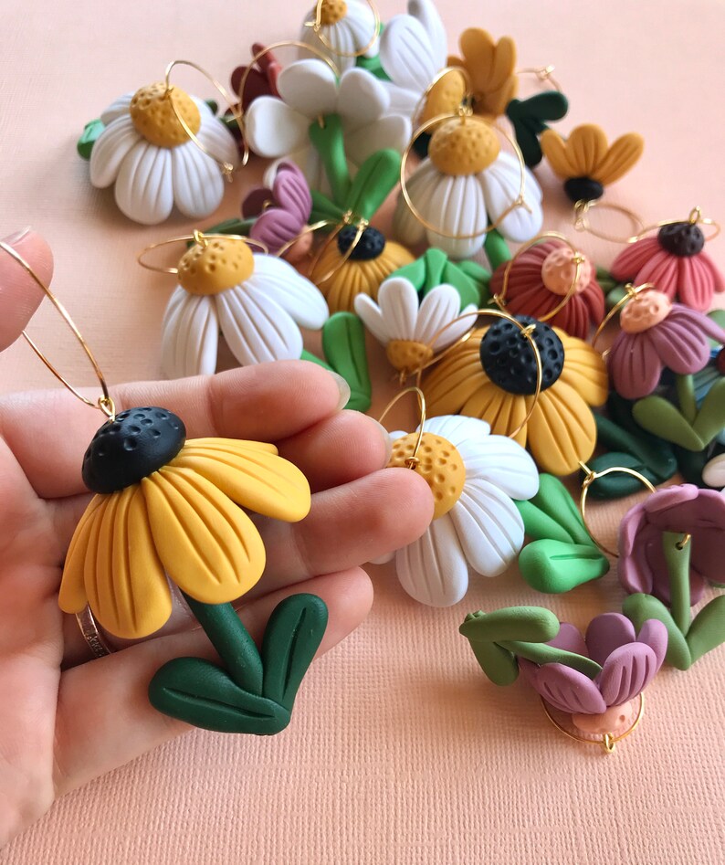 May include: A collection of handmade polymer clay flower earrings. The earrings feature a variety of flower designs, including daisies, sunflowers, and other wildflowers. The flowers are all hand-painted with vibrant colors and have a realistic appearance.
